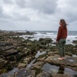Femme contemplant le littoral breton depuis des rochers de granite à marée basse, paysage côtier sauvage en Bretagne
