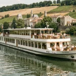 Bateau de croisière fluviale naviguant sur le Rhône avec des vignobles en terrasse et un village médiéval en arrière-plan